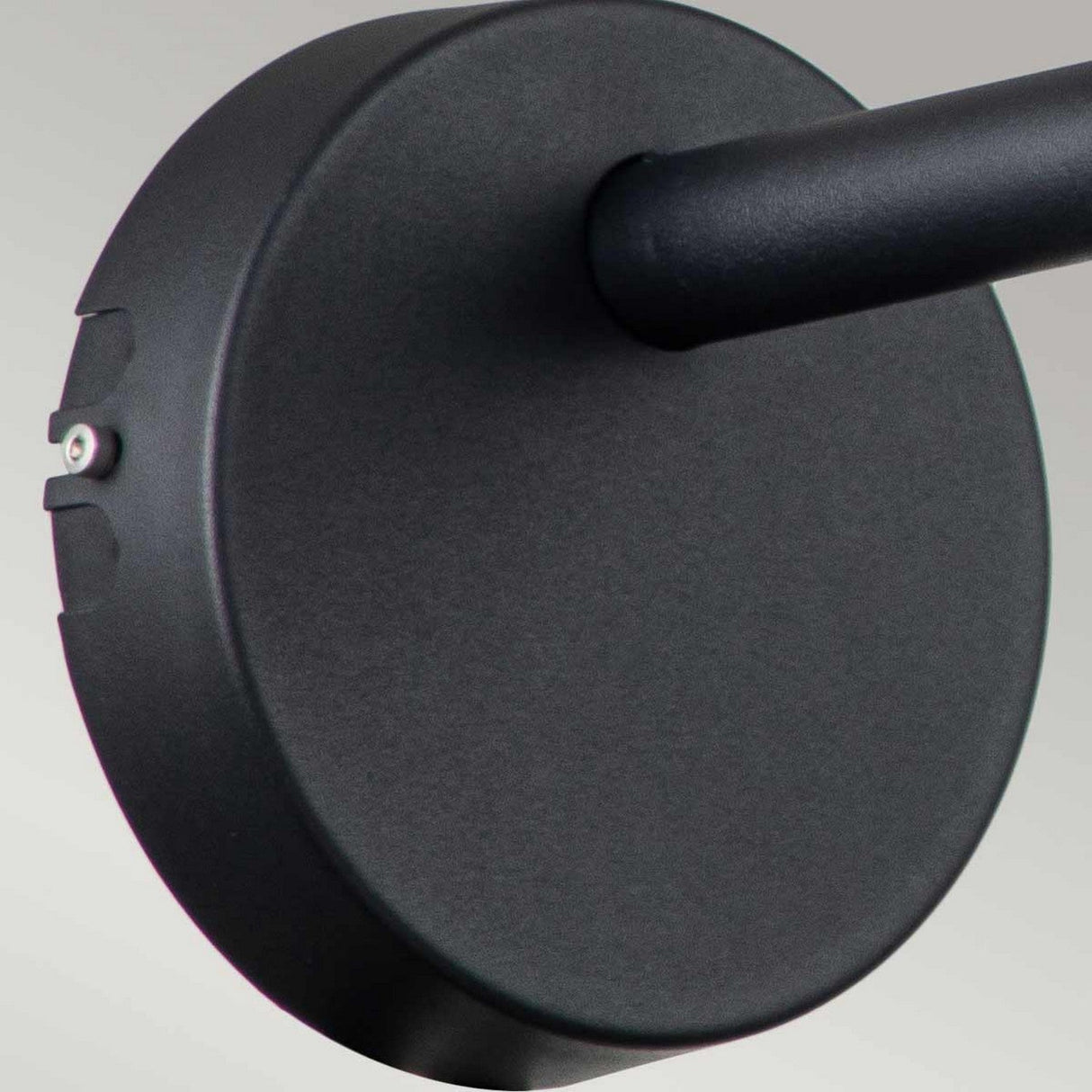 Close-up of the Selma Outdoor LED Wall Light - Black, featuring a round metal wall mount with an adjustable directional head attached to a cylindrical rod. This architectural lighting piece stands out against a neutral light gray background, showcasing its sleek design ideal for outdoor installations.