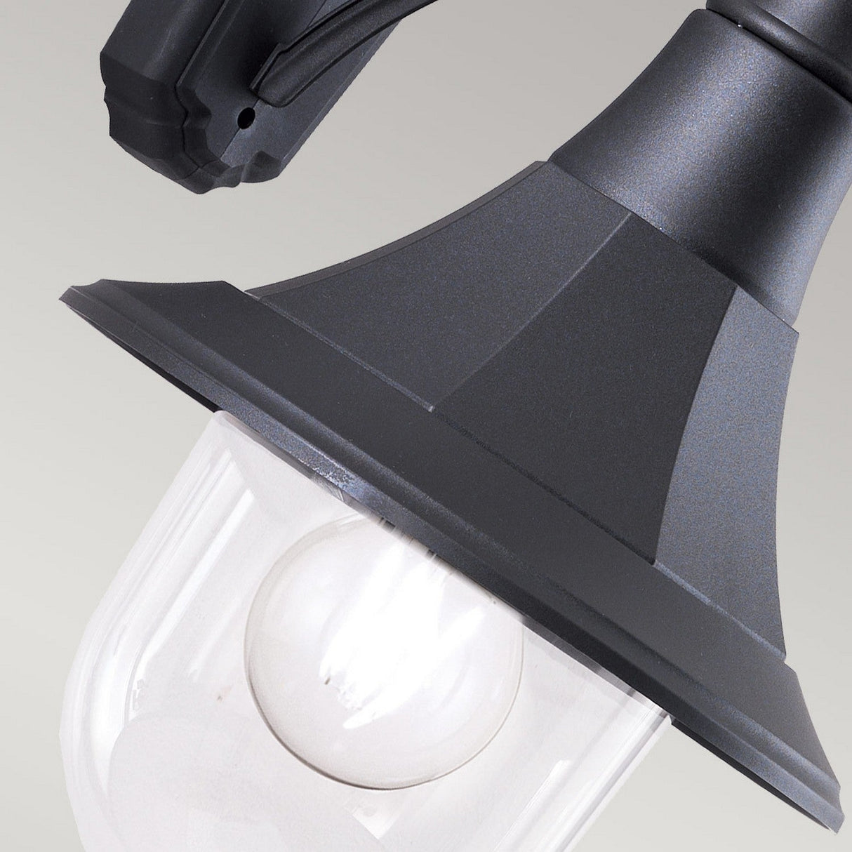 Close-up of the Shannon 1 Light Outdoor Down Wall Light in black, designed to be corrosion-proof. It features a clear glass shade that reveals a visible light bulb inside. The lantern boasts a curved, hexagonal design and an IP44 rating, complete with a mounting bracket at the top. The background is plain and light gray.