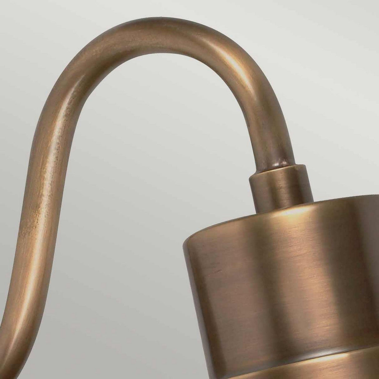 A close-up of the Sheldon 1 Light Outdoor Wall Light - Aged Brass, featuring a curved arm and a cylindrical shade against a light gray background. The brushed finish on the aged brass adds an industrial charm and a vintage appearance.