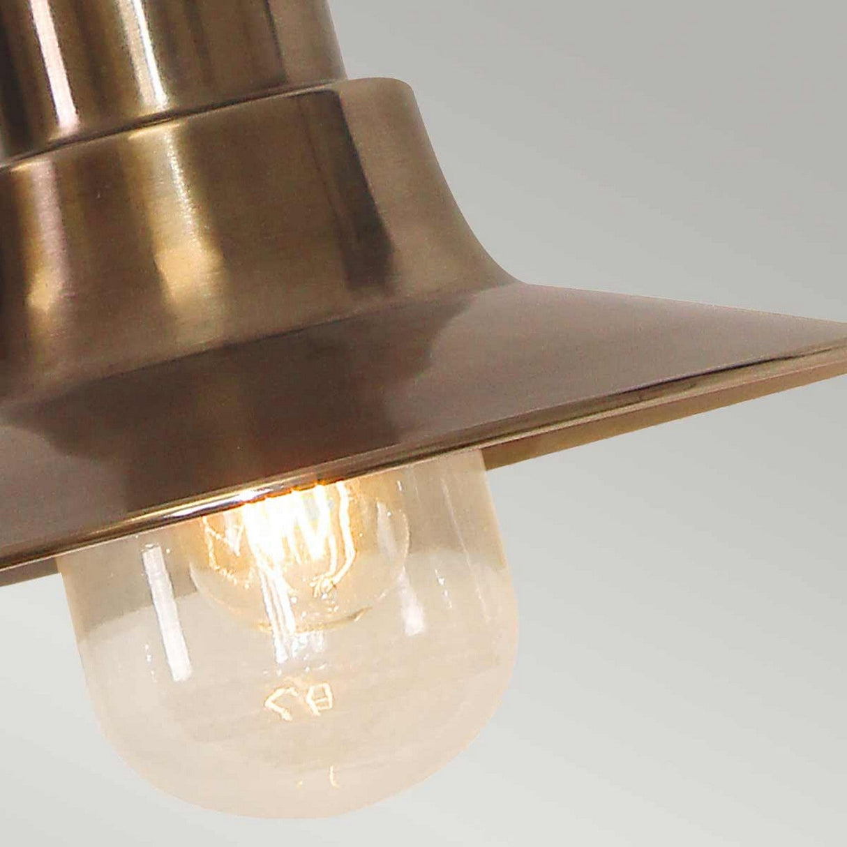 A detailed view of the Sheldon 1 Light Outdoor Chain Pendant in aged brass showcases a clear glass bulb that emits a warm glow, creating a cozy atmosphere. This vintage-inspired lamp, with its wide conical shade, is ideal for rustic outdoor lighting and is presented against a neutral backdrop.
