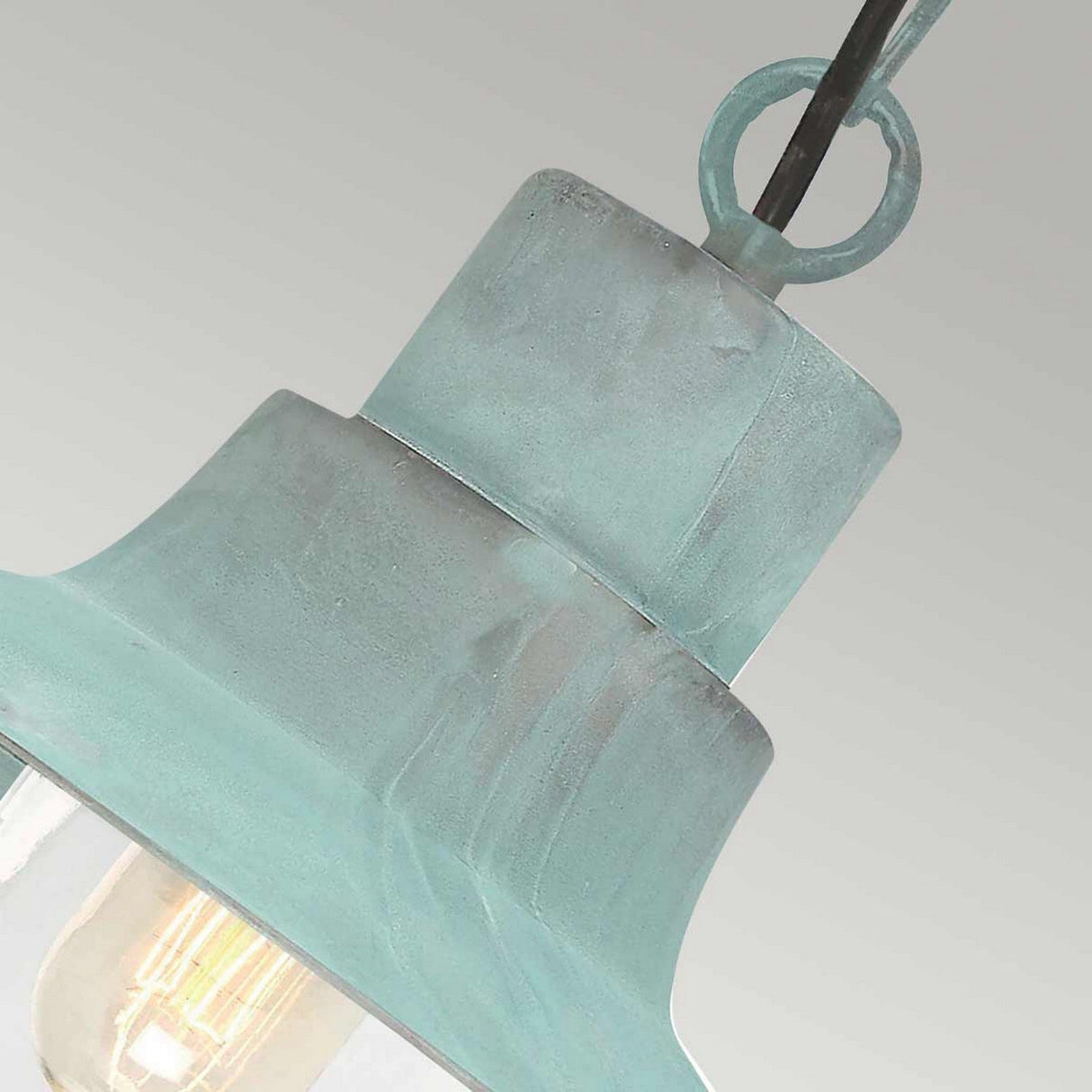 Close-up of the Sheldon 1 Light Outdoor Chain Pendant in verdigris, featuring a weathered blue-green finish and a visible filament bulb. The design includes a ring attachment at the top, embodying a vintage industrial aesthetic against a light gray background.