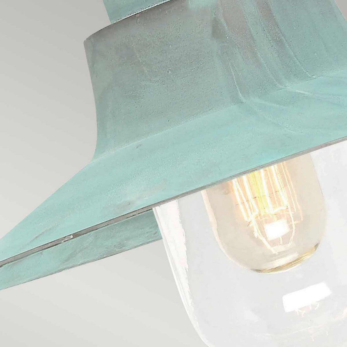 Close-up of the Sheldon 1 Light Outdoor Wall Light - Verdigris, showcasing its vintage-style design with a verdigris finish and a large, clear glass bulb beneath a wide, weathered teal metal shade. The bulb emits a warm glow against a soft gray background.