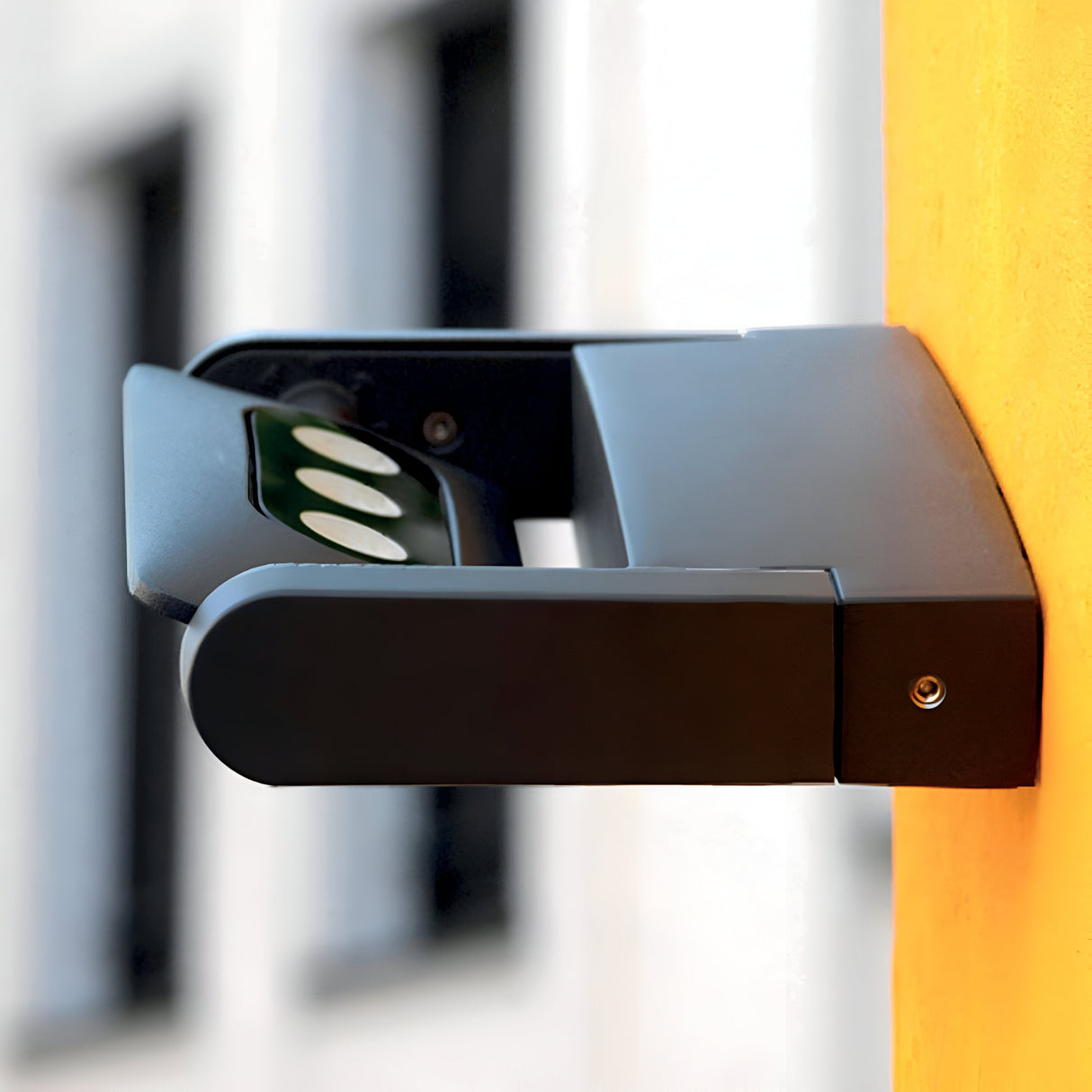 A modern graphite Sigmund 3 Light Outdoor Wall Light, featuring an IP65 rating for weather resistance, is mounted on an orange wall. In the blurred background, windows and a white building facade add contrast to the contemporary setup.