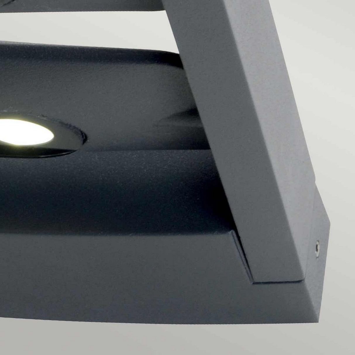 Close-up of the Sigmund 6 Light Outdoor Wall Light in a modern rectangular shape and graphite color, featuring LED technology with a glowing circular bulb. Mounted on a light gray wall, its sleek design showcases clean lines and metallic surfaces while providing an IP65 rating for durability.