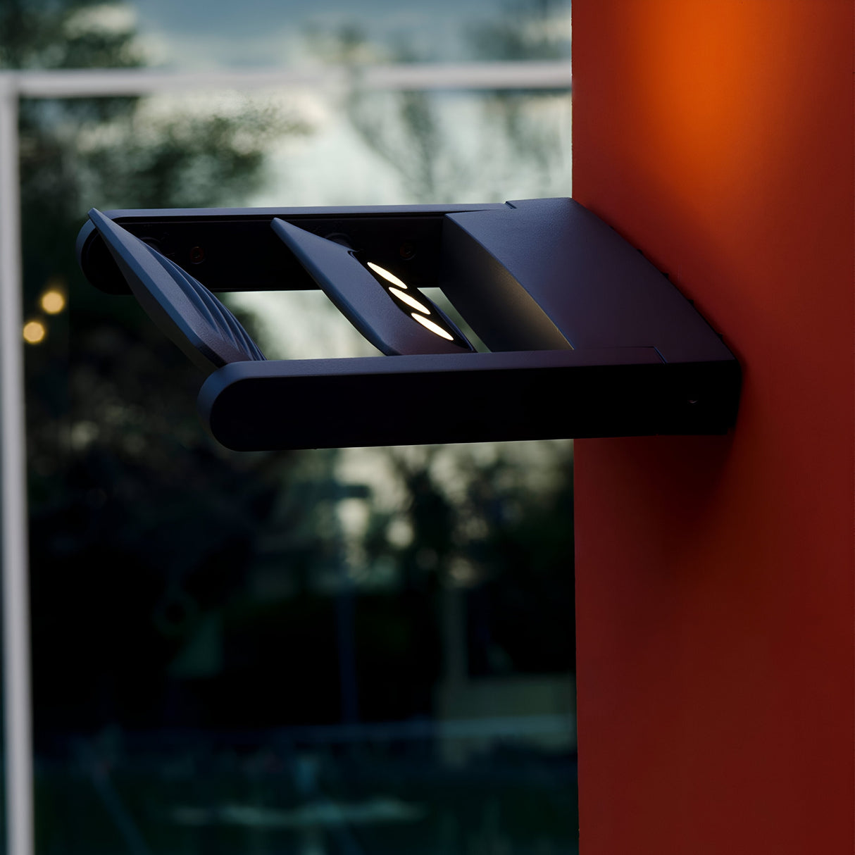 The Sigmund 6 Light Outdoor Wall Light in graphite is installed on an orange wall. Its contemporary rectangular design boasts a sleek black finish and utilizes energy-efficient LED technology to cast light downward, illuminating both the wall and the surrounding area. In the background, blurred foliage and reflective glass enhance the setting.