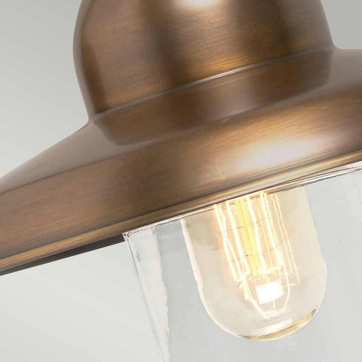Close-up of the Somerton 1 Light Outdoor Wall Light in aged brass, featuring an exposed filament bulb within a clear glass enclosure. The soft illumination accentuates the warm tones of the fixture.