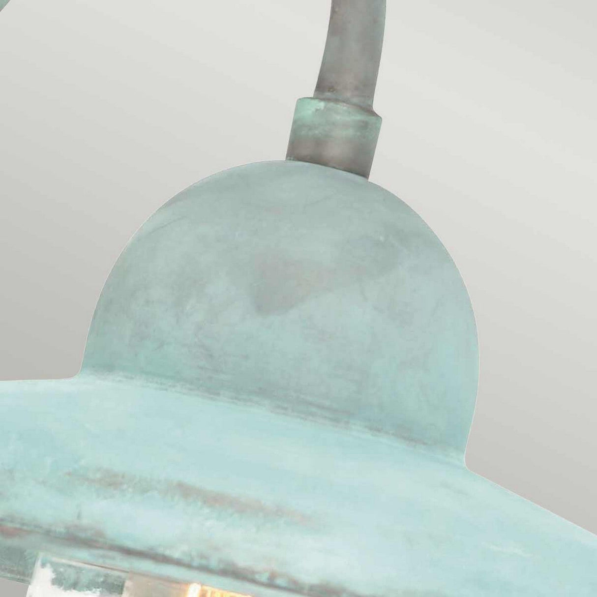 A detailed view of the Somerton 1 Light Outdoor Wall Light showcases its rustic, weathered design featuring a dome-shaped top and a curved arm. The verdigris finish provides a greenish-blue hue that enhances its vintage charm, set against a simple gray background that emphasizes its elegant design.