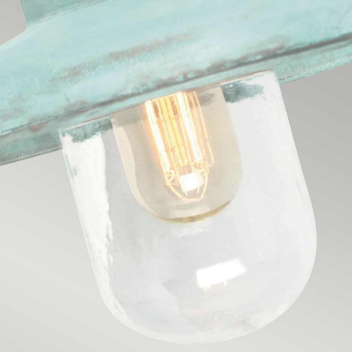 A close-up view features the Somerton 1 Light Outdoor Wall Light with its vintage-style bulb encased in a clear glass fixture, highlighted by a teal metallic verdigris finish. The softly glowing filament of the bulb creates a warm ambiance, reminiscent of an outdoor wall light against a gradient of light gray in the background.