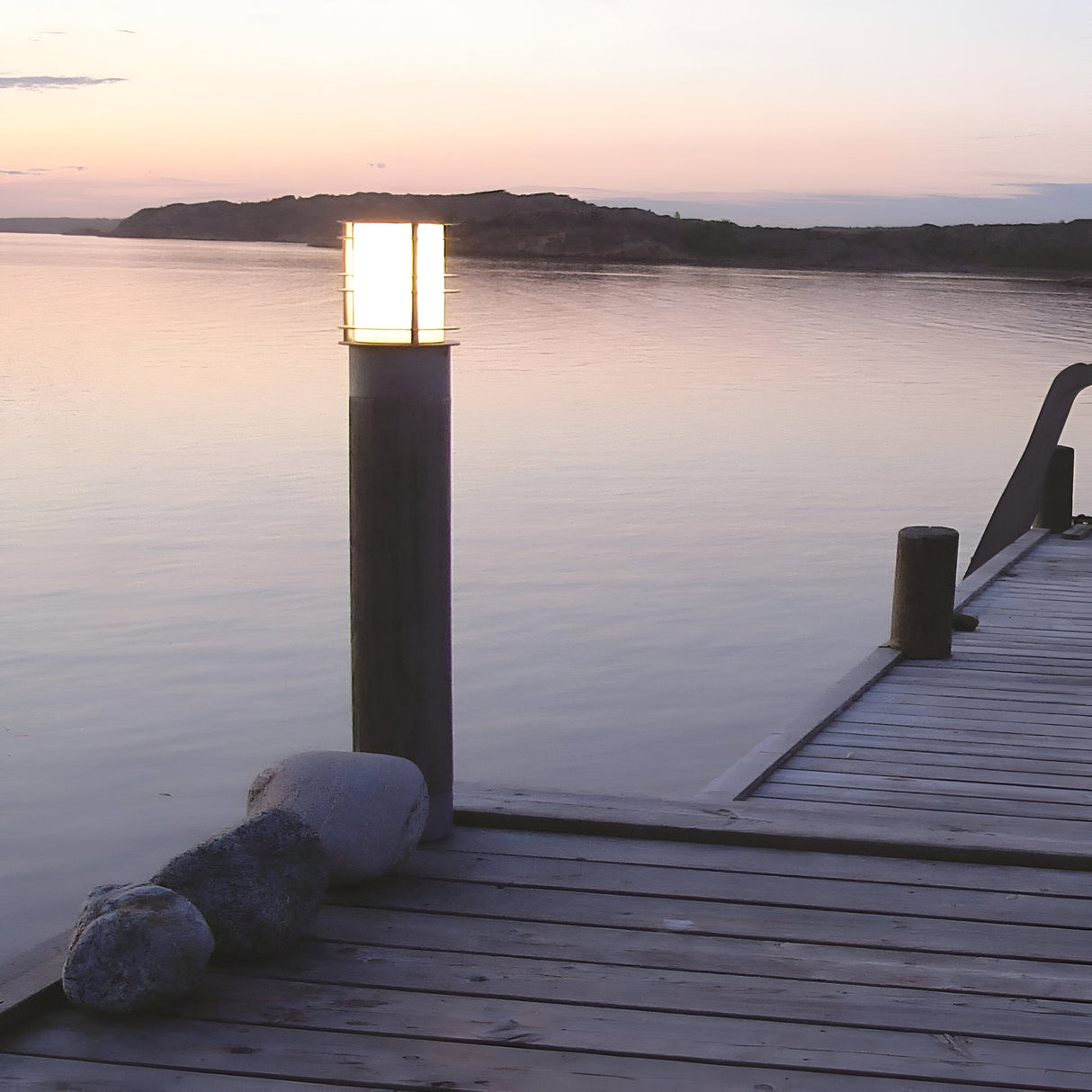 A tranquil sunset view from a wooden dock extending over calm water features outdoor lighting with the Stockholm 1 Light Large Bollard - Black casting a warm glow. The sky is painted with soft pastel hues, hills are visible in the distance, and stones are placed near a large galvanized steel bollard at the dock's edge.