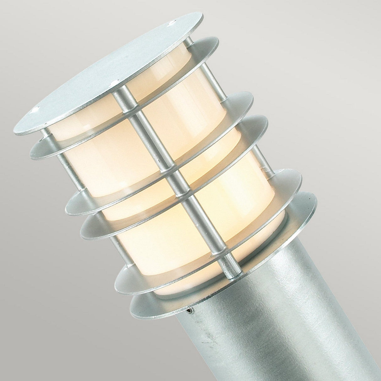 A close-up view of the Stockholm Large Bollard - Silver highlights its cylindrical design featuring tiered metal rings, crafted in galvanized steel with a silver finish, encircling a glowing cream-colored light. The sleek metal structure strikingly contrasts with the soft illumination.