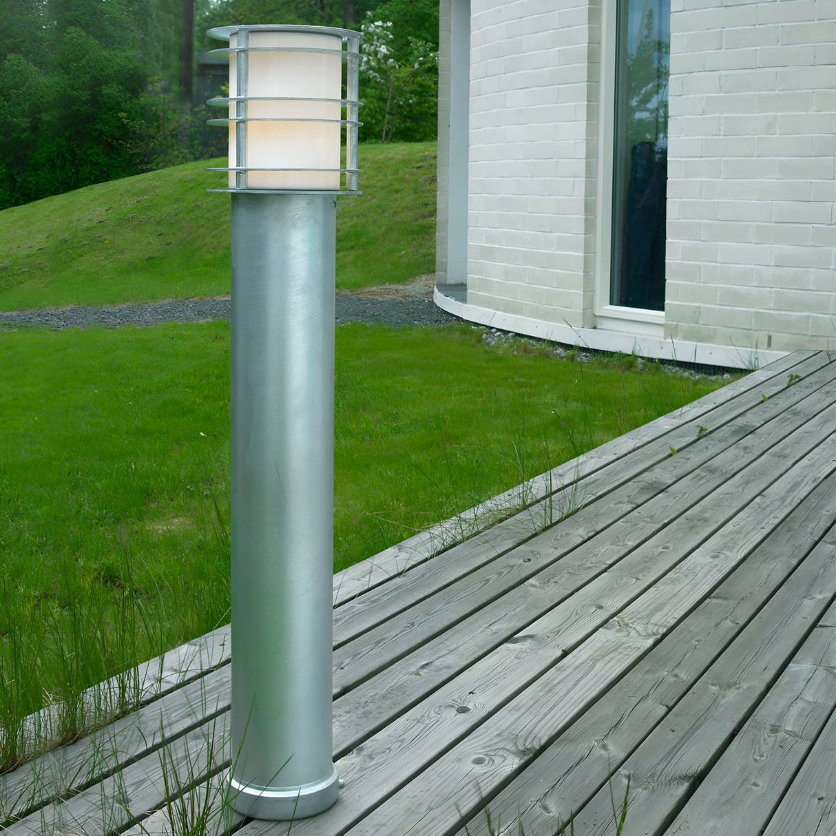 A contemporary, cylindrical outdoor lamp crafted from galvanized steel stands on a wooden deck adjacent to a building with white brick walls. This illuminated Stockholm Large Bollard - Silver elegantly lights up the area. In the background, a lush green grassy landscape enhances the tranquil setting.