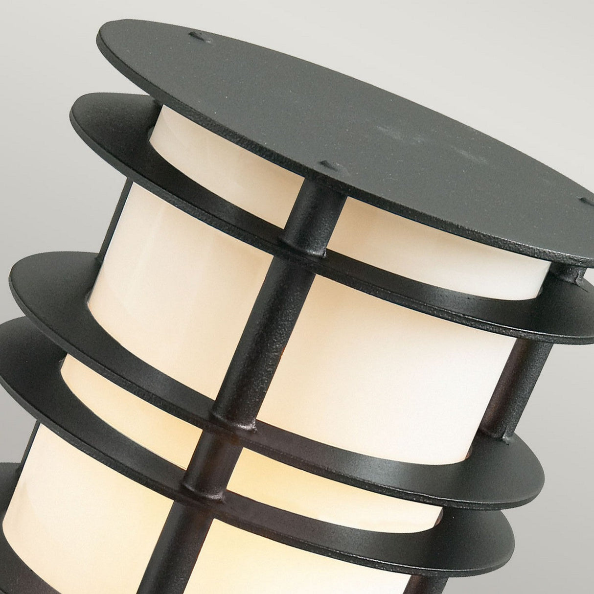 A close-up of the Stockholm 1 Light Small Bollard - Black reveals its modern Scandinavian style with a cylindrical shape. It showcases white panels and black metal bands that form a layered design. Constructed from galvanized steel, this light emits a soft glow against a neutral background.
