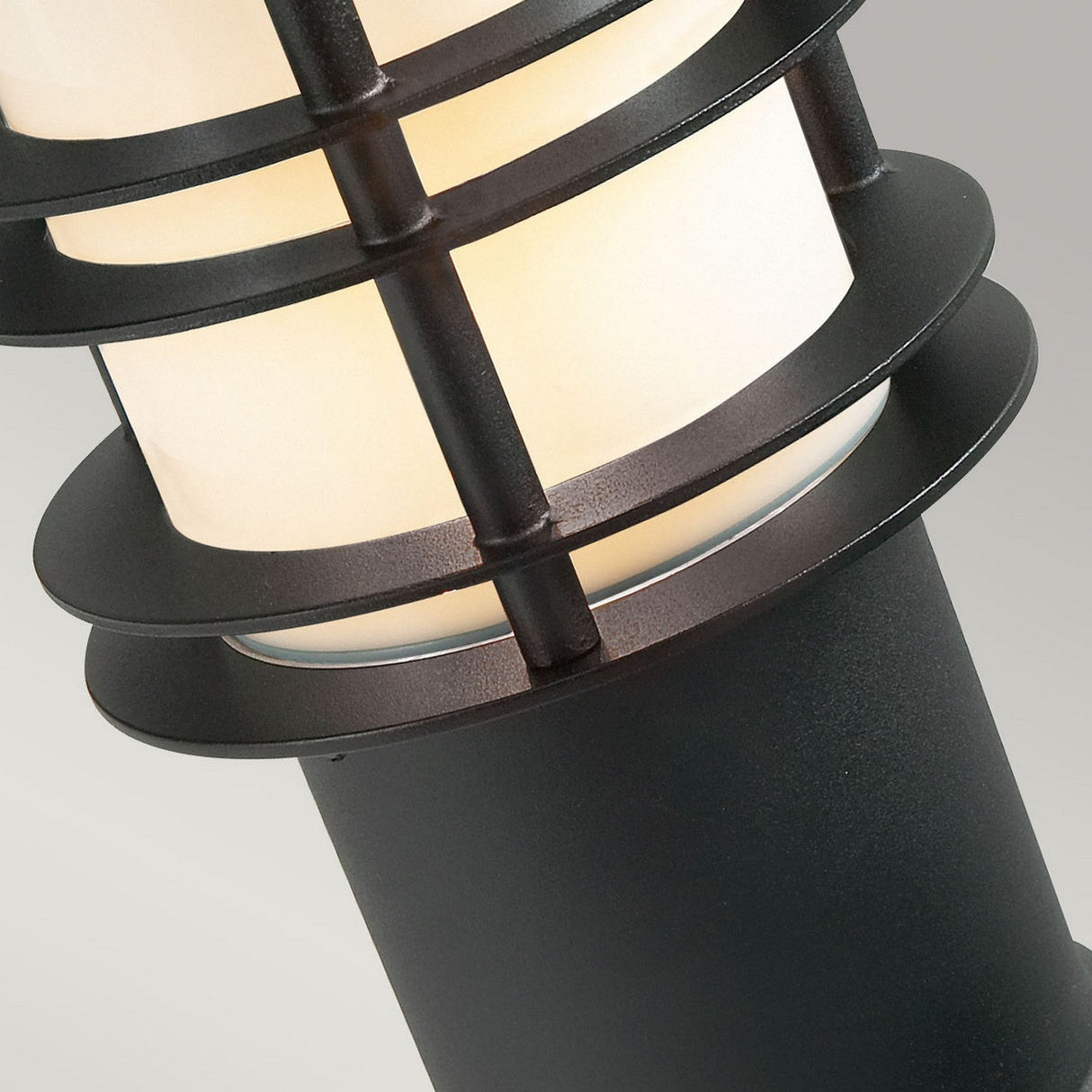 Close-up of the Stockholm 1 Light Small Bollard in a Scandinavian style, highlighting its cylindrical white lamp within a sleek black frame adorned with horizontal rings. The neutral gray background emphasizes its modern design.