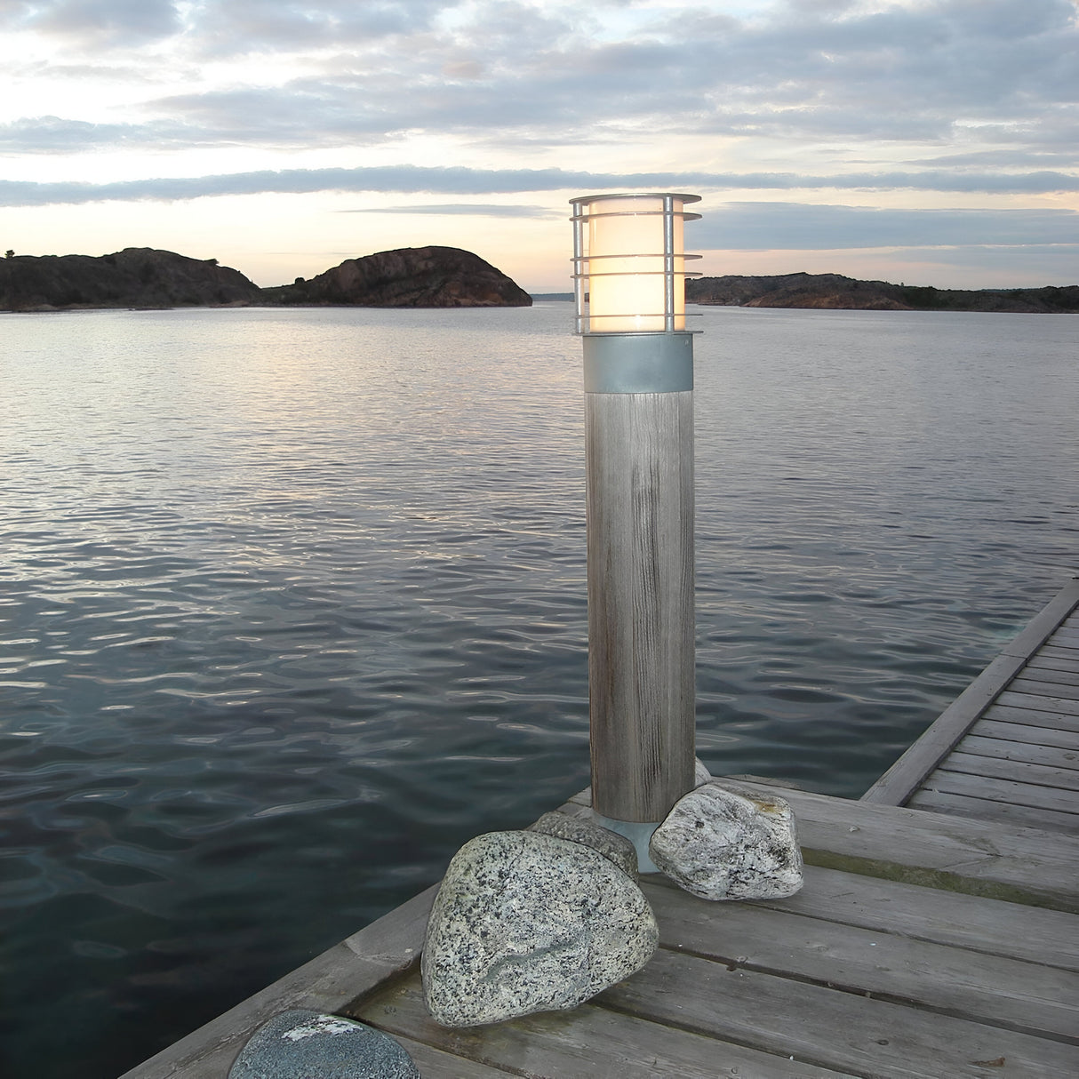 The Stockholm Large Bollard in Natural Wood and Silver, crafted with a modern design, stands on a wooden pier surrounded by stones. This IP65-rated bollard emits a warm glow. In the background, calm waters and rocky formations lie under a cloudy sky at dusk.