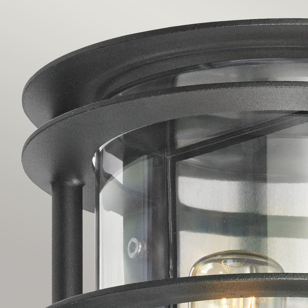 Close-up of the Stockholm Outdoor 1 Light Flush Light, featuring a black metal frame and clear glass panes that reveal the bulb inside. The cylindrical design exudes an industrial aesthetic against a soft gradient of light gray background.