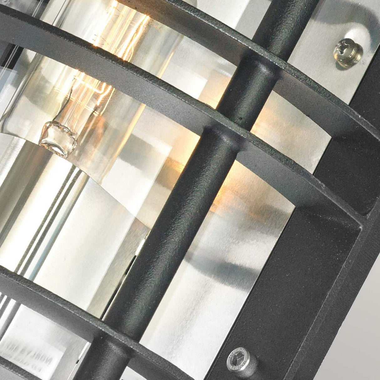 Close-up of the Stockholm Outdoor 1 Light Flush Light - Black With Clear Glass, showcasing its industrial-style design with a visible filament bulb encased in a sleek frame. The warm glow beautifully illuminates the metallic surface, enhancing its industrial aesthetic.