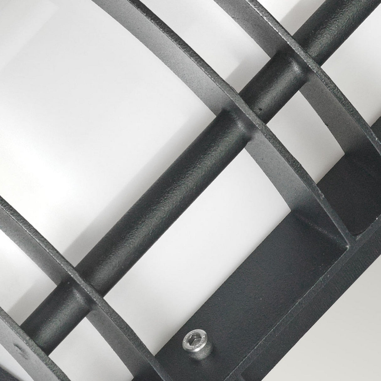 Close-up of the Stockholm Outdoor 1 Light Flush Light in black, featuring a corrosion-resistant metal grid structure set against a smooth white background. The design includes intersecting vertical and horizontal bars with a small screw positioned at one of the intersections.