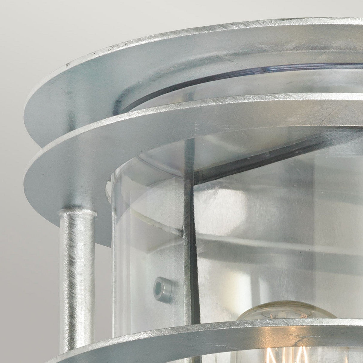 Close-up of the Stockholm Outdoor 1 Light Flush Light in silver with a cylindrical design. It features a clear glass enclosure and a galvanized steel frame, visible are support rods and a light bulb inside. This IP54 rated fixture offers sleek and industrial metallic finishes for any stylish space.