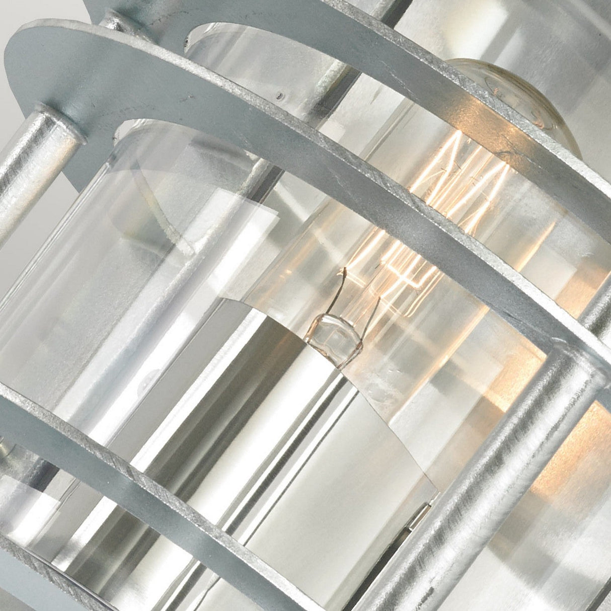 A close-up of the Stockholm Outdoor 1 Light Flush Light in silver, showcasing its modern industrial design with galvanized steel and clear glass panels. The fixture features a visible glowing filament bulb inside, complemented by metallic bars surrounding the bulb. With an IP54 rating, it offers a sleek and contemporary style ideal for exterior spaces.