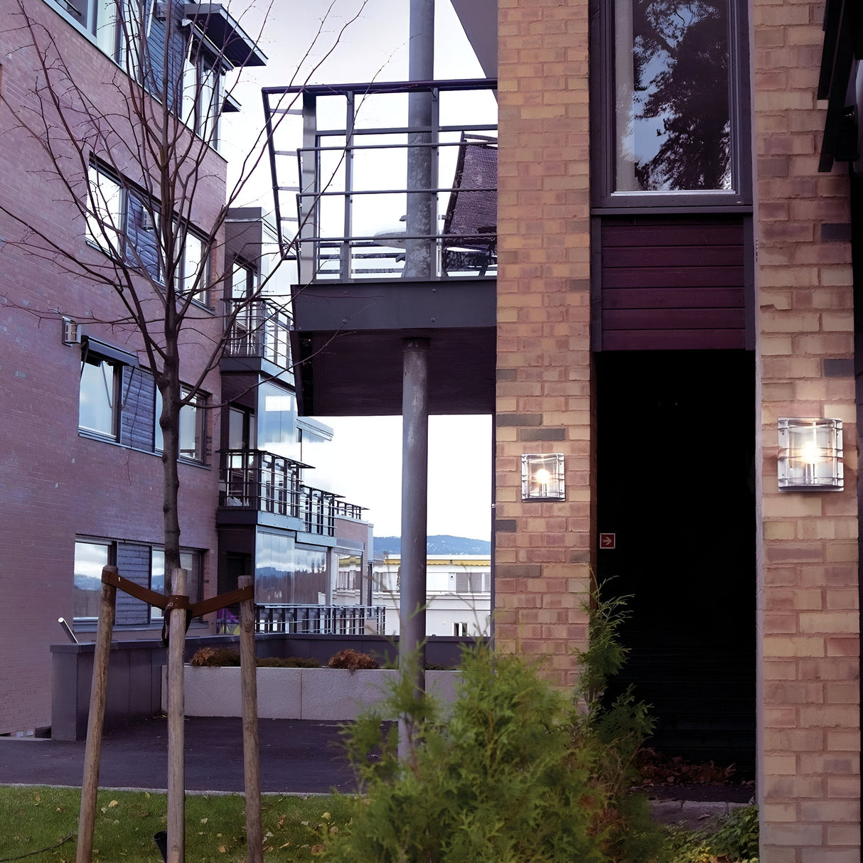 A view of modern apartment buildings with brick exteriors is separated by a narrow path lined with bare trees and shrubs. The scene is subtly illuminated by a Stockholm Outdoor 1 Light Flush Light in silver with clear glass, which is IP54 rated. The sky is overcast, and a distant body of water peeks between the structures.