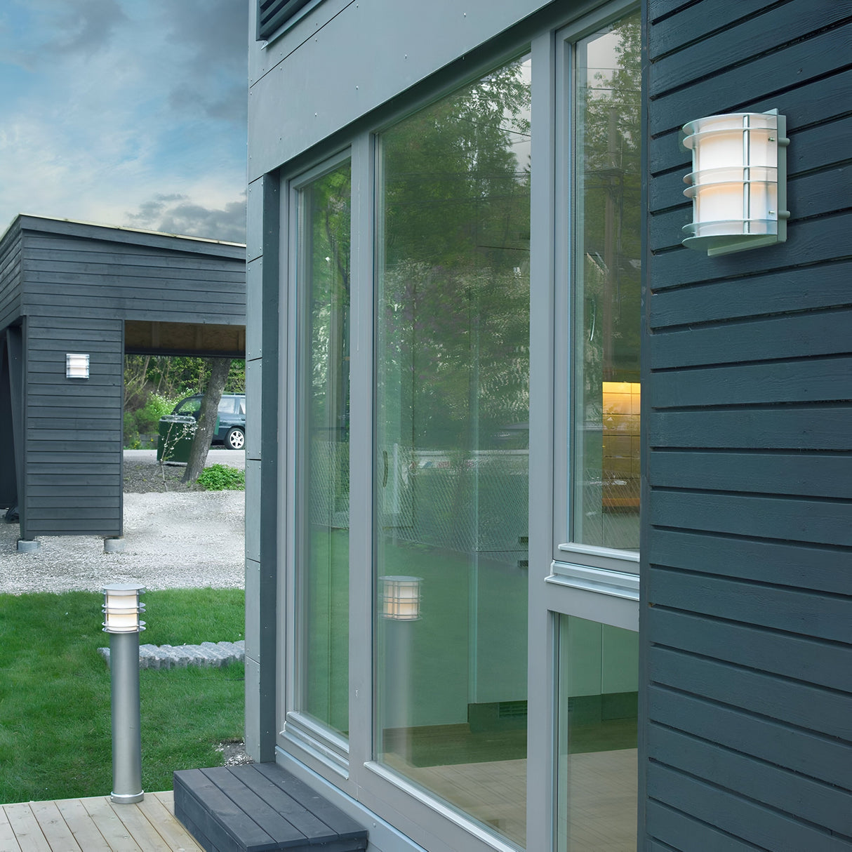 The modern house exterior showcases large glass sliding doors that reflect the surrounding greenery. Dark wooden siding is complemented by a minimalist Stockholm Outdoor 1 Light Flush Light in silver with opal glass. In the background, a driveway leads to a garage, all encompassed by the tranquility of a well-maintained lawn.