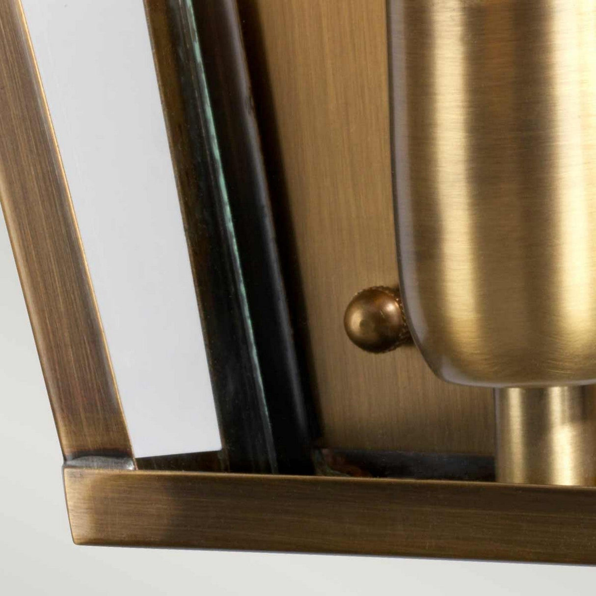 A close-up of the St James 1 Light Outdoor Wall Light reveals its British design, highlighted by a rectangular aged brass frame and a glass panel. The surface displays a brushed texture, and there is a small, spherical accent present.