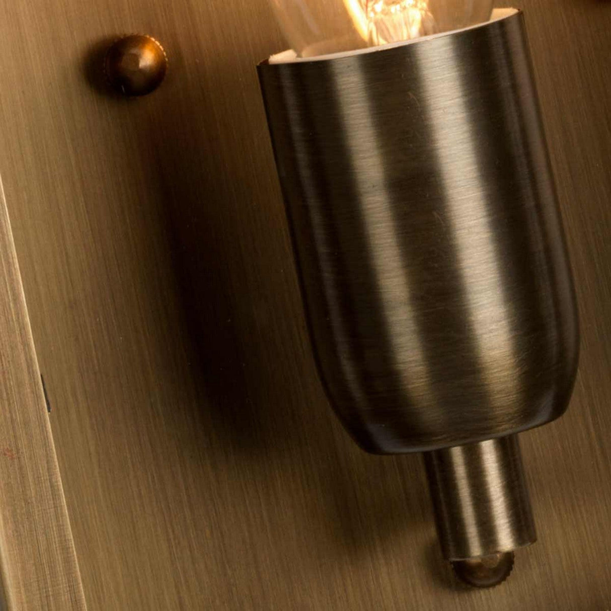 Close-up of the St Martins 1 Light Outdoor Wall Light featuring a hand-crafted, vintage-style bulb with a dark metallic holder, mounted on an aged brass wall plate. The illuminated bulb casts a warm glow on the surface.