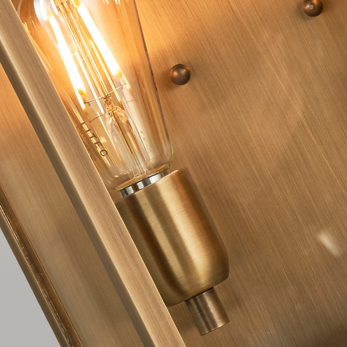 A detailed view of the St Martins 2 Light Outdoor Large Wall Light in aged brass, showcasing its vintage-style Edison bulbs emitting a warm glow. This fixture is mounted on a textured, brushed metal surface adorned with visible rivets, exuding historical charm with its warm, industrial tone.