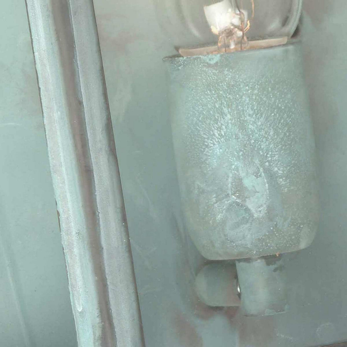 Close-up of a frosted light bulb partially covered with dust and grime, nestled next to the textured verdigris finish of the St Martins 1 Light Outdoor Wall Light, enhancing its industrial, weathered appearance.