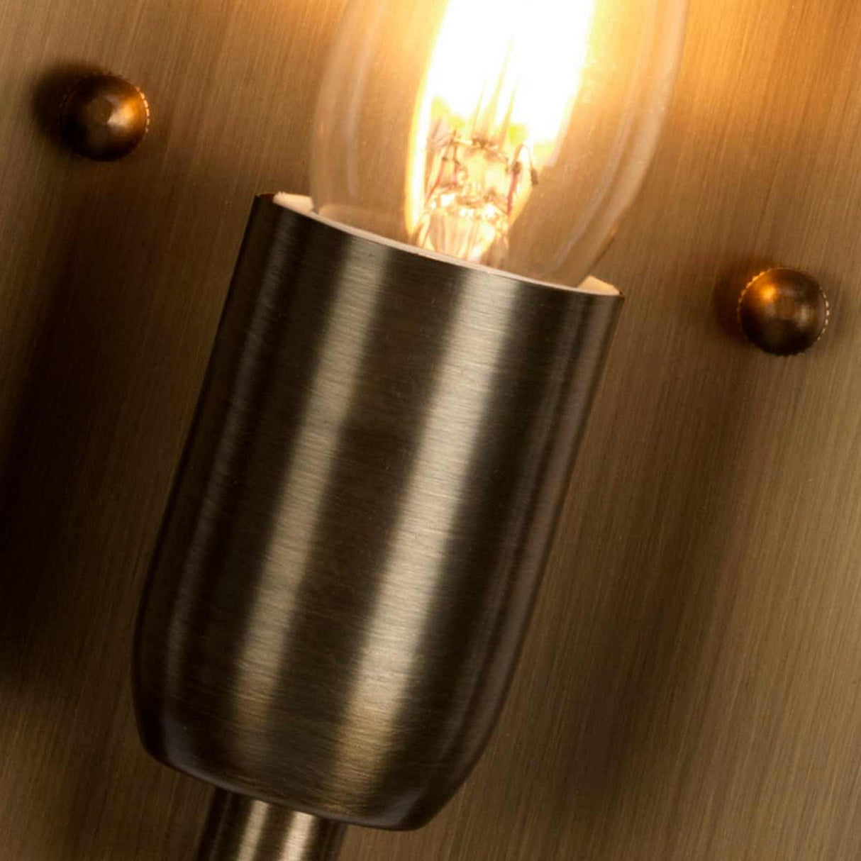 Close-up of a lit bulb in the St Pauls 1 Light Outdoor Wall Light - Aged Brass, mounted on a textured surface. A warm glow highlights the bulb's filament and holder details, while two brass screws are visible beside the fixture, enhancing its rustic charm.