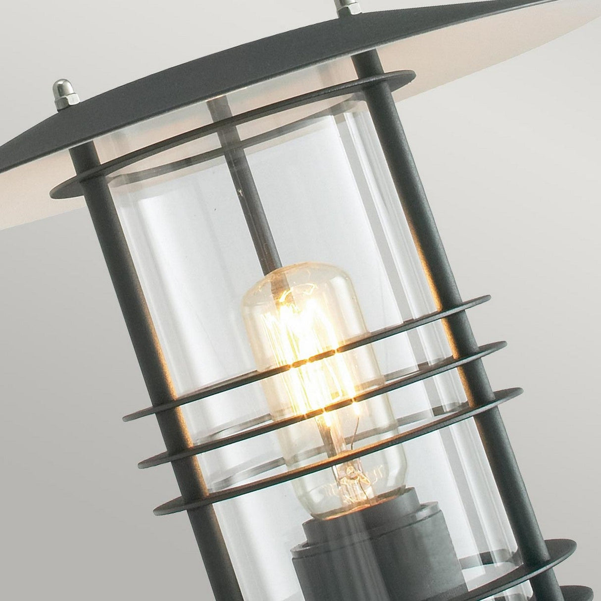 A close-up of the Stockholm Outdoor 1 Light Wall Lantern in black showcases its modern design with a clear glass casing. The cylindrical lamp, rated IP54 for durability, emits a warm glow. Its sleek appearance is enhanced by horizontal bars encasing the bulb, complementing any exterior space beautifully.