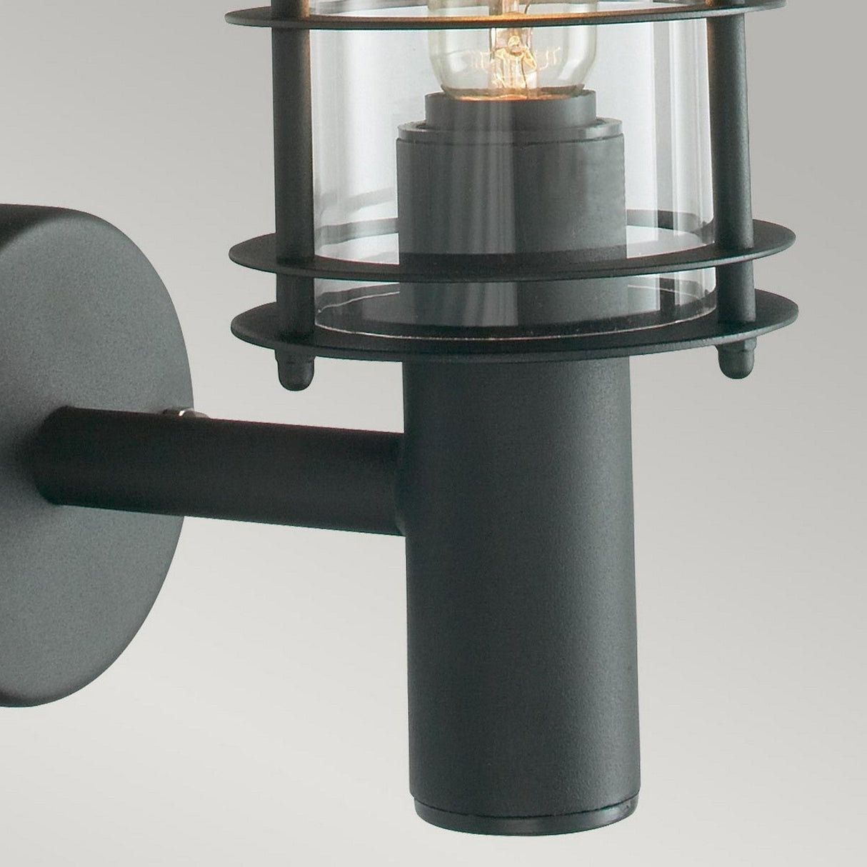 Close-up of the Stockholm Outdoor 1 Light Wall Lantern in black, featuring a sleek cylindrical metal frame and a clear glass shade with an IP54 rating. The visible lamp casts a warm glow against the gray background, seamlessly combining durability and style.
