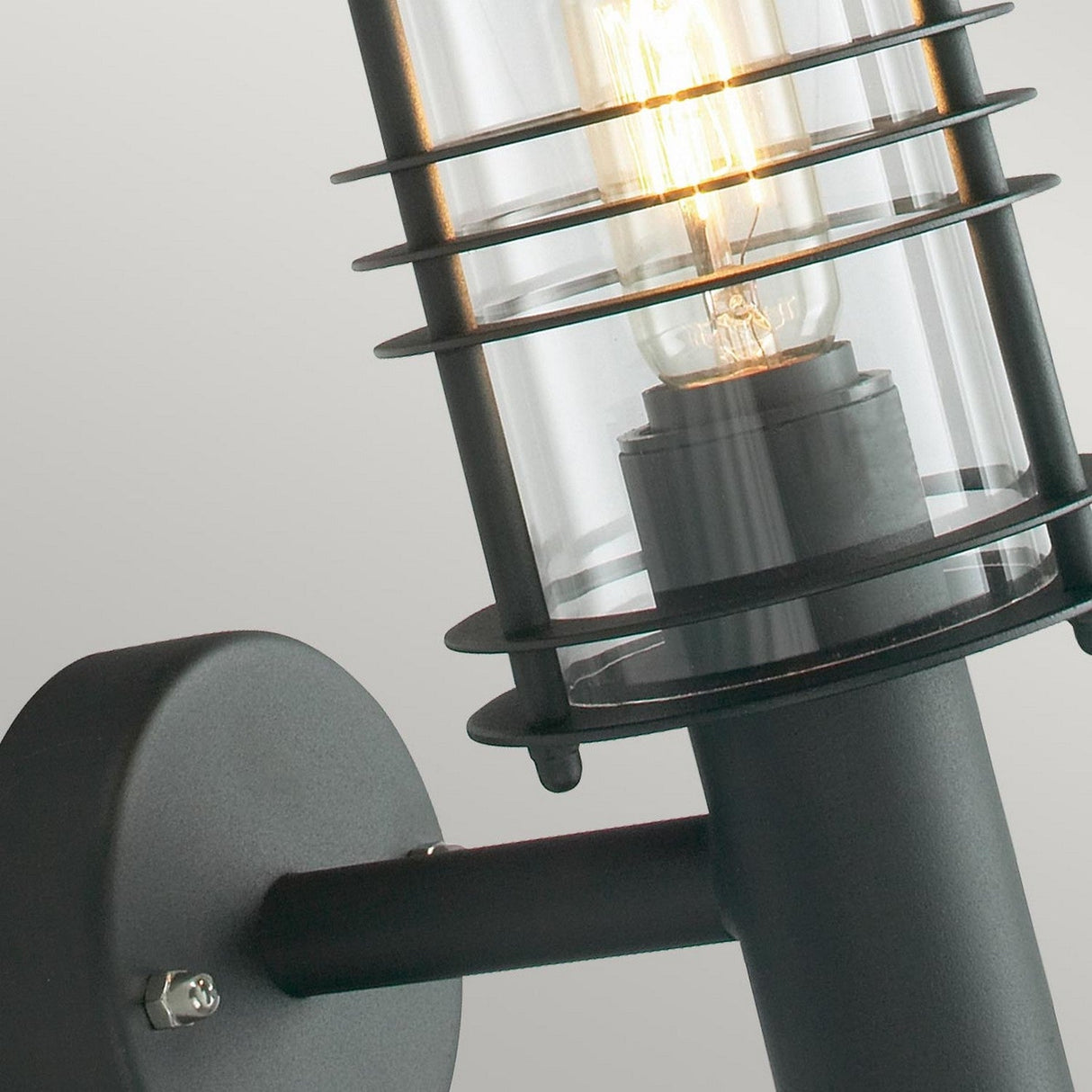 This close-up showcases the Stockholm Outdoor 1 Light Wall Lantern - Black, with its black-finished metal and clear glass cover, highlighting an exposed vintage-style light bulb. Mounted on a round base, it serves as a perfect outdoor wall lantern set against a plain light gray backdrop.