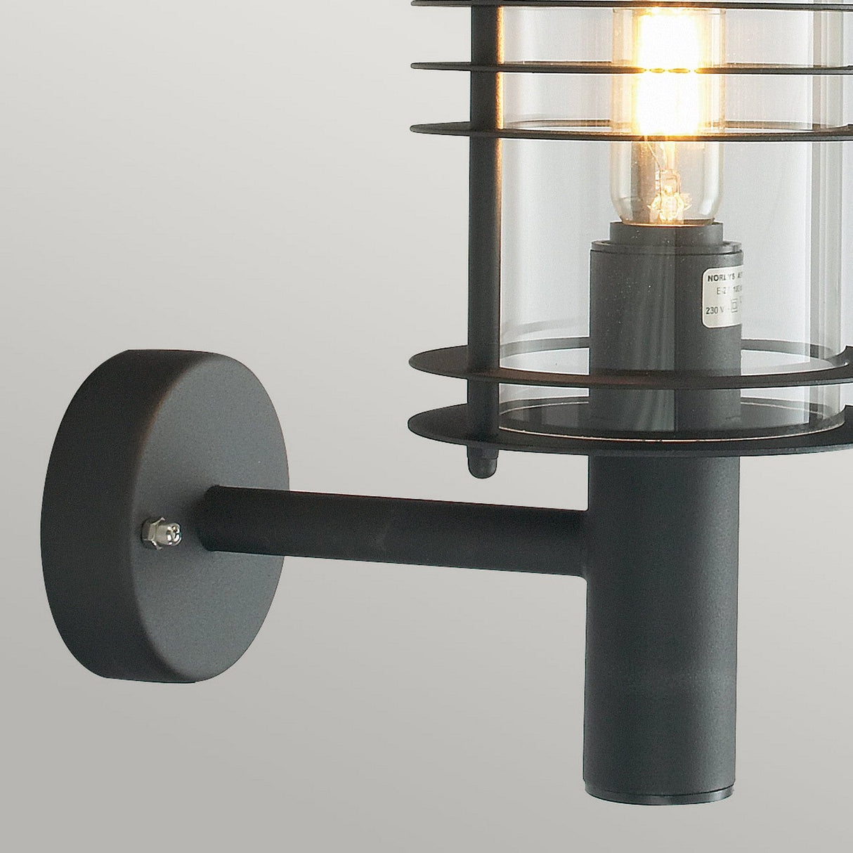 The Stockholm Outdoor 1 Light Large Wall Lantern in black is a wall-mounted fixture featuring a cylindrical glass enclosure with a visible light bulb inside. Its modern design and durable structure are secured to a circular wall mount with a short horizontal arm, providing sleek elegance to any space.