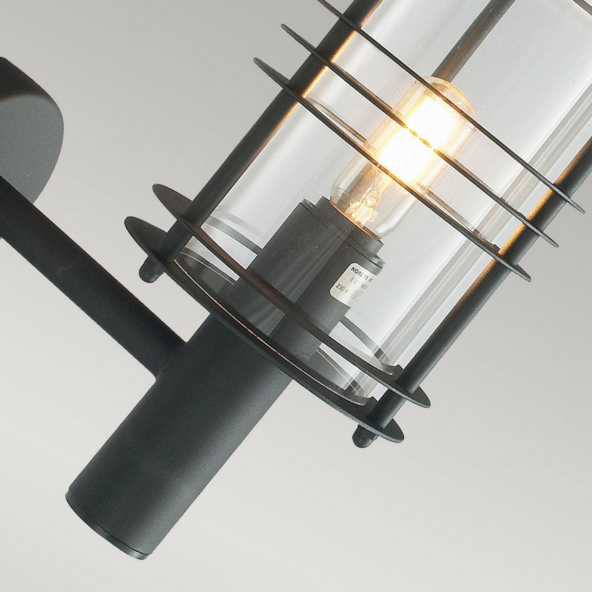 Close-up of the Stockholm Outdoor 1 Light Large Wall Lantern, showcasing its modern industrial design with a black metal frame surrounding a cylindrical clear glass casing. Inside, a lit incandescent bulb emits a warm glow. This durable lighting fixture seamlessly blends with the neutral gray background, epitomizing modern design.
