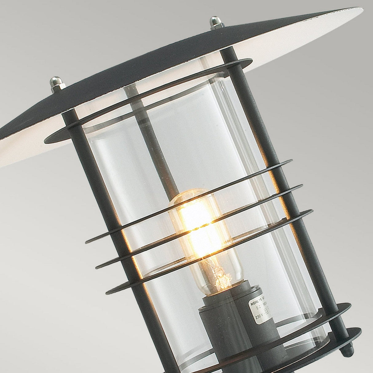 The image showcases a close-up of the Stockholm Outdoor 1 Light Large Wall Lantern in black, which consists of a sleek metal frame surrounding a glass cylinder. Within, a light bulb shines brightly, epitomizing contemporary design. The background displays a delicate gray gradient.