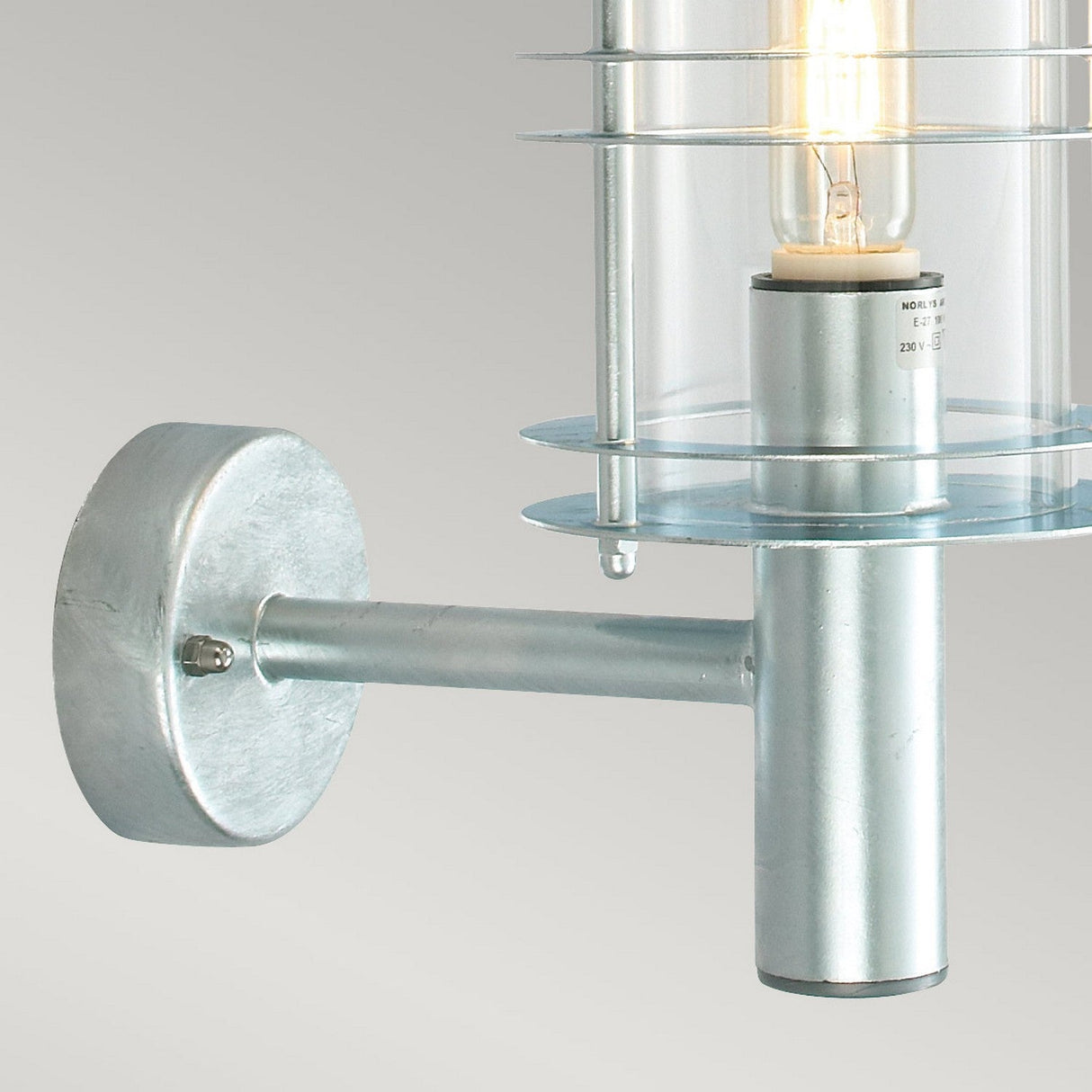 Close-up of the Stockholm Outdoor 1 Light Large Wall Lantern in silver, featuring a cylindrical galvanized steel base and a clear glass shade. The visible bulb highlights its sleek, industrial design, making it ideal for outdoor lighting.
