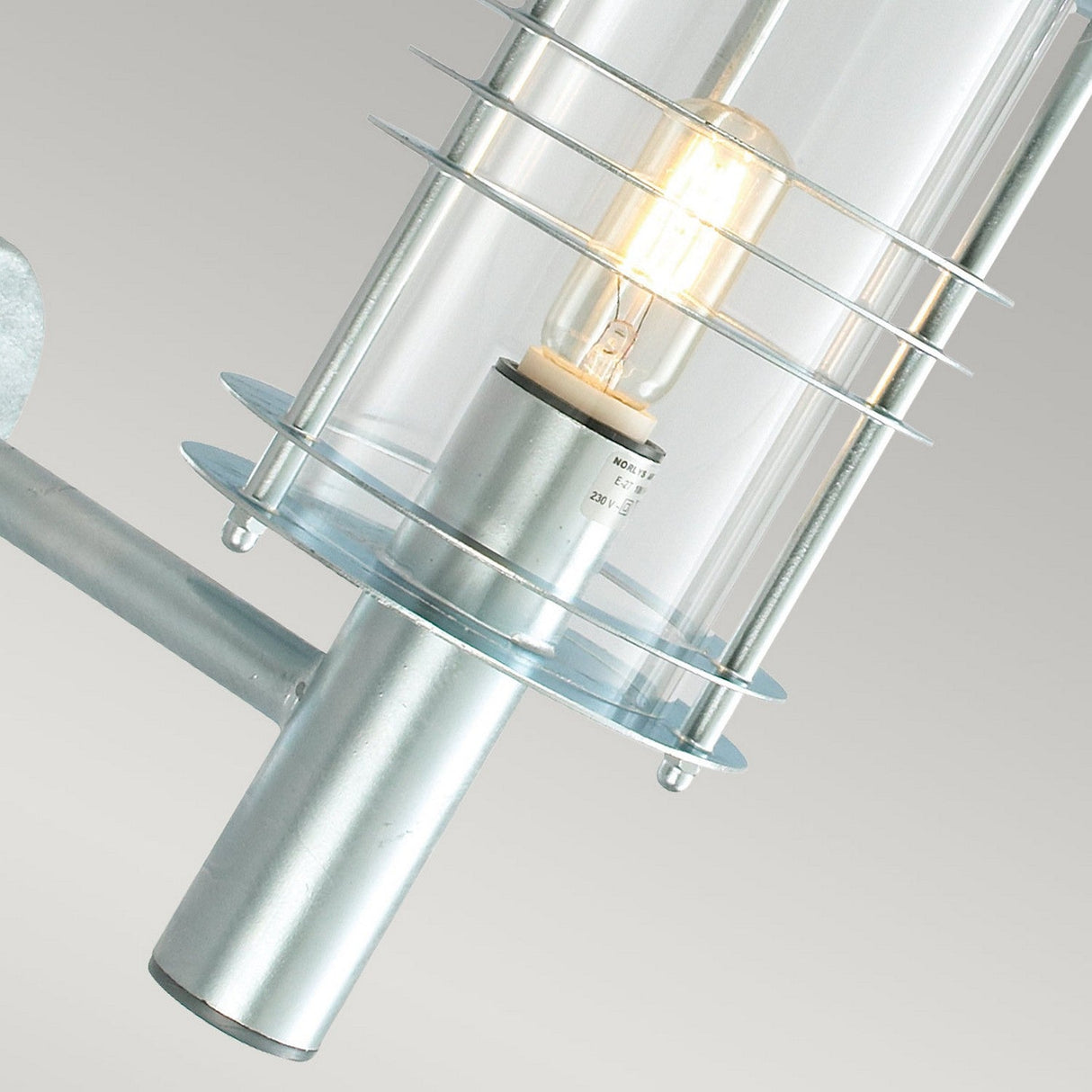 Close-up of the Stockholm Outdoor 1 Light Large Wall Lantern in a modern cylindrical design, crafted from galvanized steel. The bulb is encased in clear glass with metal accents and mounted on an elegant silver base. Boasting an IP54 rating, it withstands the elements. The plain background enhances its focus.