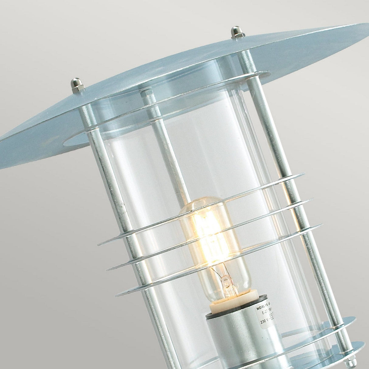 Close-up of the Stockholm Outdoor 1 Light Large Wall Lantern in a modern cylindrical design, crafted from galvanized steel with a silver finish. It showcases a clear glass casing and metal accents, with an illuminated bulb inside. Rated IP54 for durability and elegance, it stands out beautifully against a plain, light gray background.