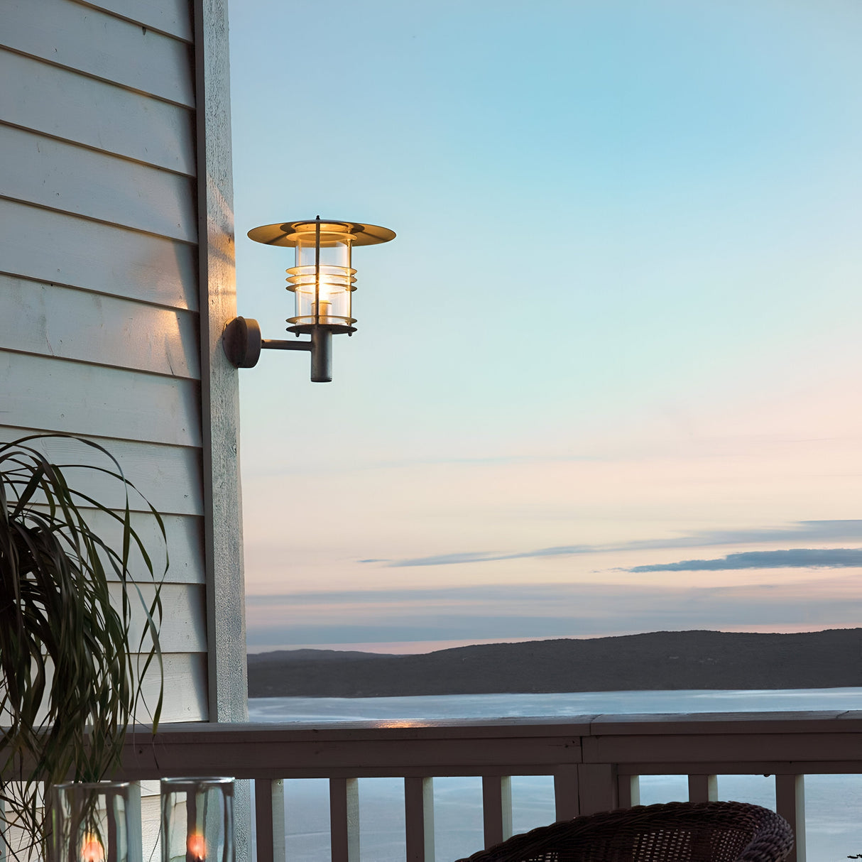 The Stockholm Outdoor 1 Light Large Wall Lantern in silver casts a warm glow on the scenic view of the ocean and distant hills at dusk, framed by soft pastel-colored skies. Made from galvanized steel with an IP54 rating, it is designed to withstand the elements. A plant is partially visible on the left side of this picturesque scene.