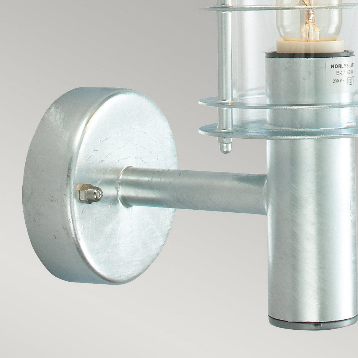 A close-up of the Stockholm Outdoor 1 Light Wall Lantern - Silver features its cylindrical glass cover enclosing a light bulb. The IP54-rated silver fixture, made from galvanized steel, is attached to a round wall plate and set against a light gray background.