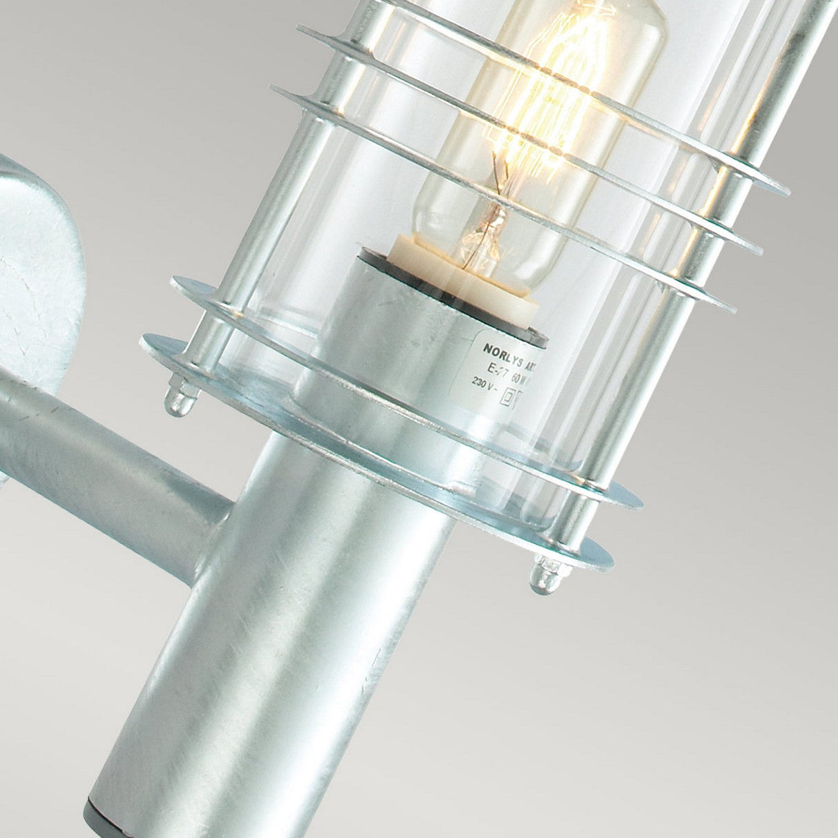 Here is an updated sentence using the specified product data:
Close-up of the Stockholm Outdoor 1 Light Wall Lantern in silver, crafted from galvanized steel and glass, showcasing an exposed filament bulb. The lantern is illuminated, casting a warm glow and encased within a protective wire frame, making it ideal for IP54-rated outdoor wall applications.