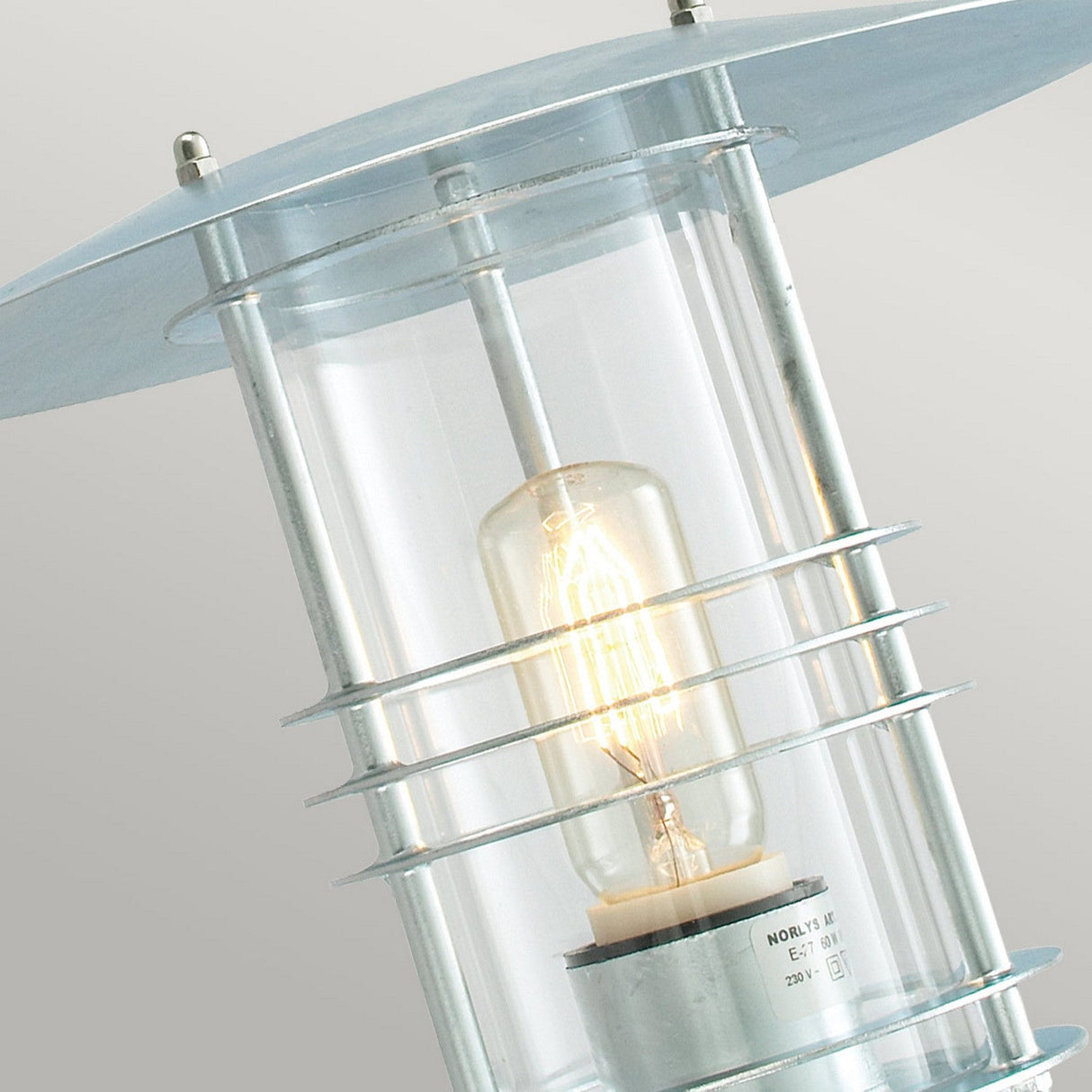 Close-up of the Stockholm Outdoor 1 Light Wall Lantern in silver featuring a visible filament bulb. The fixture showcases a galvanized steel frame with horizontal bars encircling the glass, offering a contemporary design and is IP54 rated for weather resistance.