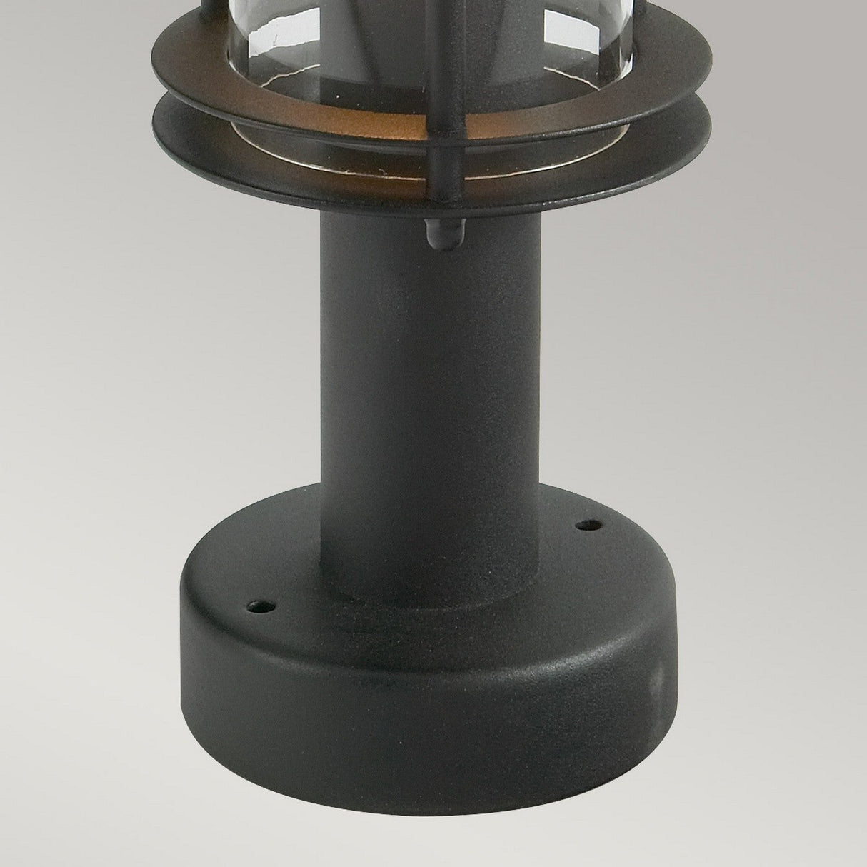 A black metal outdoor lantern, known as the Stockholm 1 Light Pedestal - Black, is made from galvanized steel and features a cylindrical base with a clear glass enclosure. Set against a light gray background, its design incorporates layered metal rings at the top, offering a modern and sleek appearance.