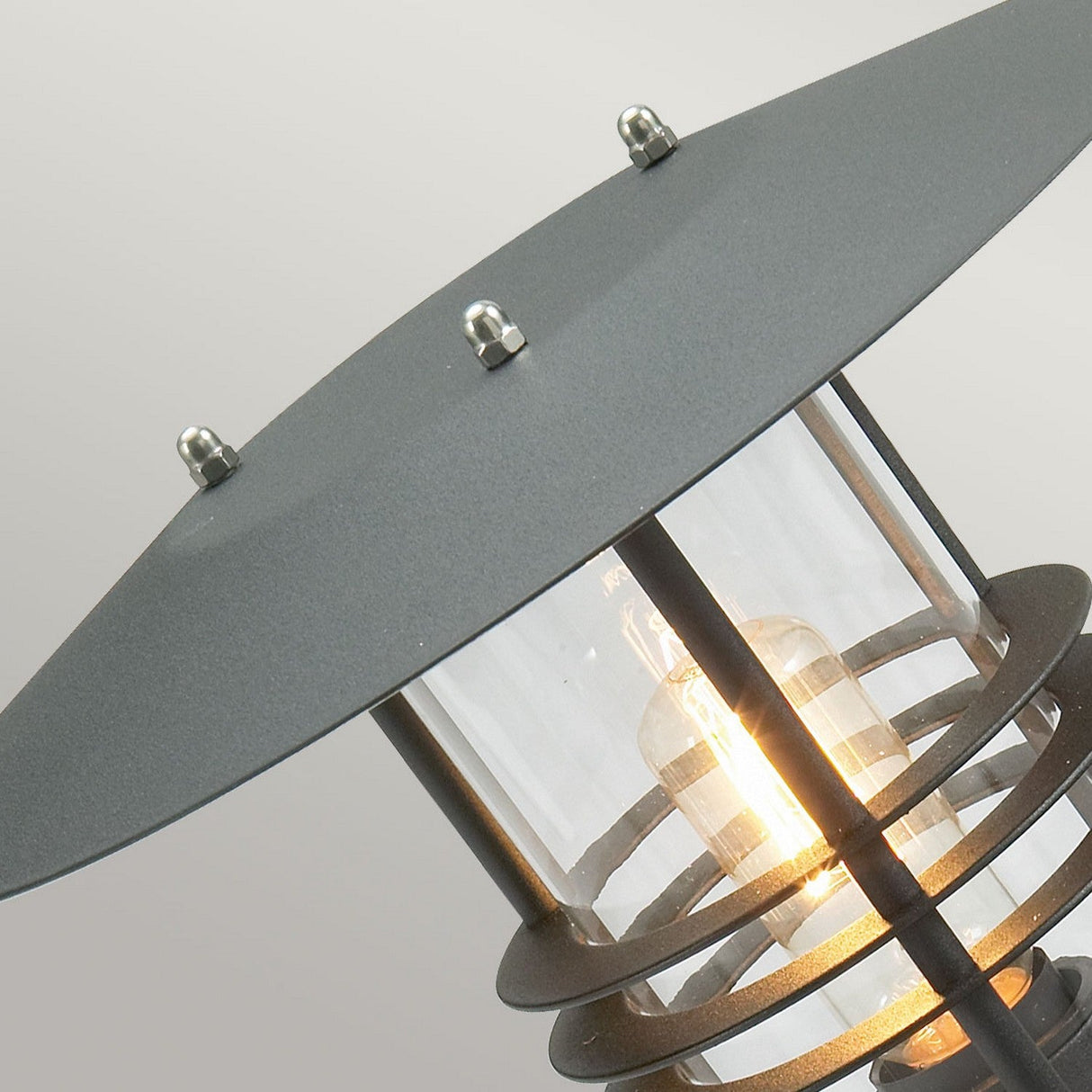 Close-up of a modern outdoor light fixture, the Stockholm 1 Light Pedestal - Black, highlighting a glowing bulb. The design features a sleek galvanized steel frame with a wide, flat circular top set against a soft gray backdrop.