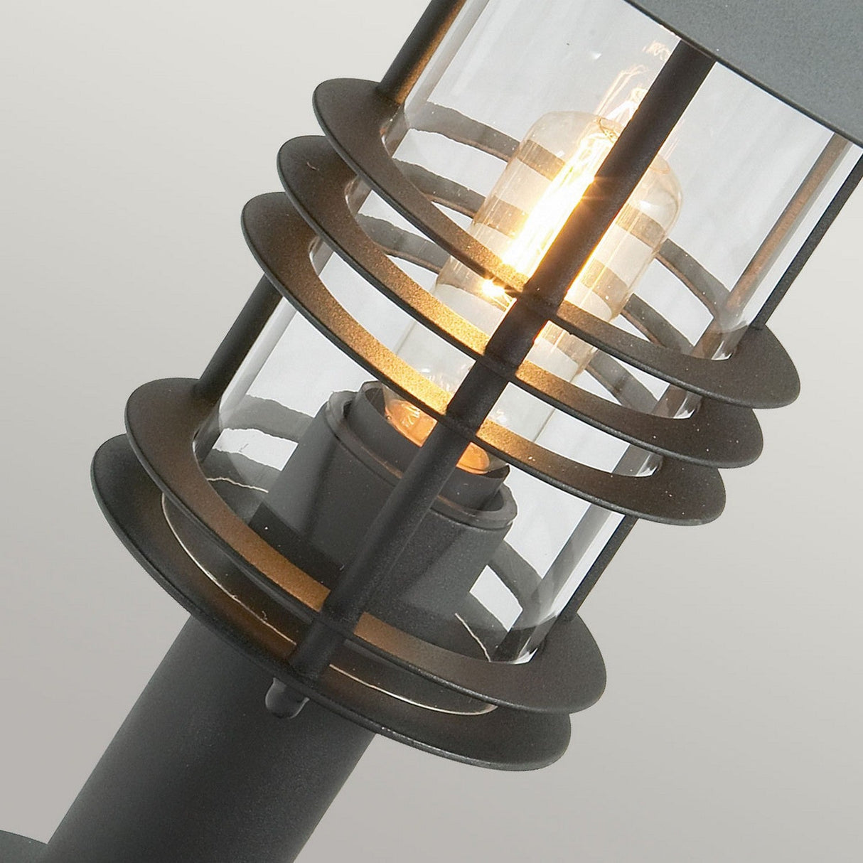 Close-up of a modern outdoor lamp with a glowing light bulb. The Stockholm 1 Light Pedestal - Black features a cylindrical glass casing surrounded by horizontal rings in galvanized steel, creating an industrial look. The background is a neutral gray, perfect for highlighting sophisticated outdoor lighting.