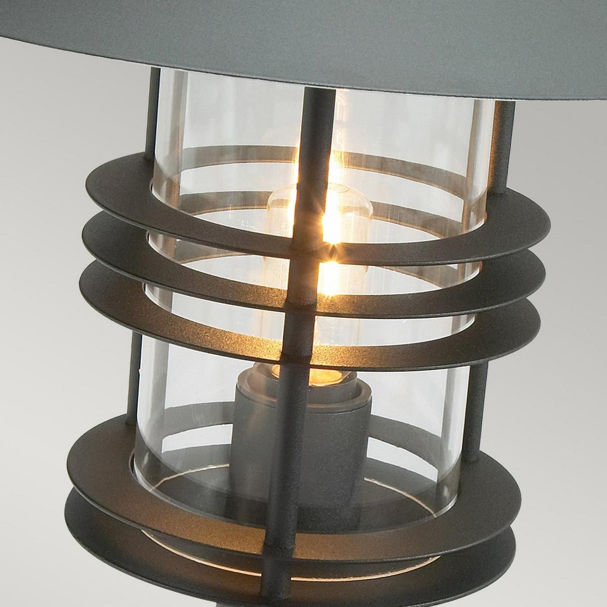 Close-up of the Stockholm 1 Light Grande Pedestal in a sleek black finish, showcasing its cylindrical glass enclosure and metal rings surrounding a glowing light bulb, set against a neutral background.