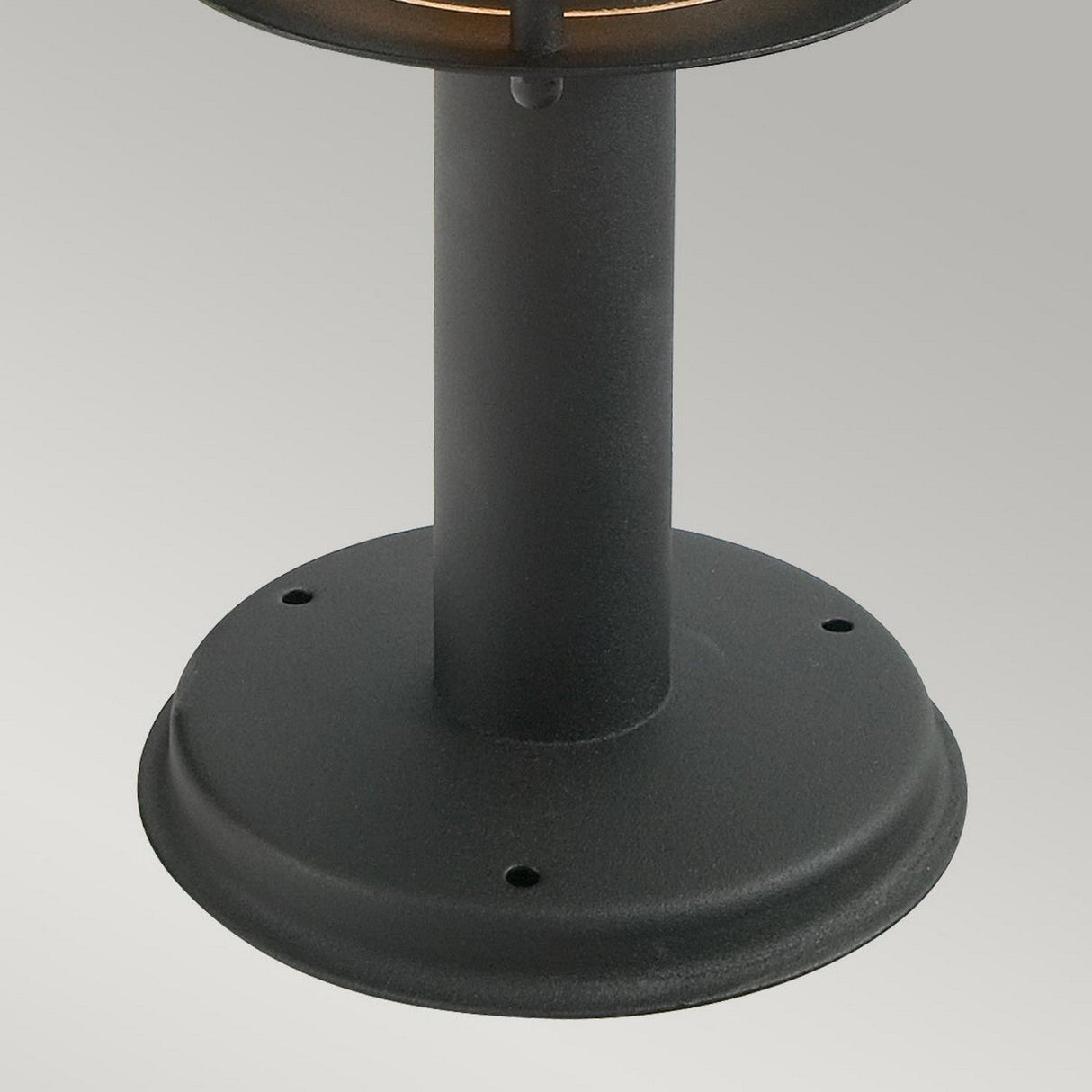 The Stockholm 1 Light Grande Pedestal - Black showcases its Scandinavian design with a close-up of its sleek black cylindrical base featuring a round, flat bottom. Crafted from galvanized steel, it includes small mounting holes and is set against a plain gray background.