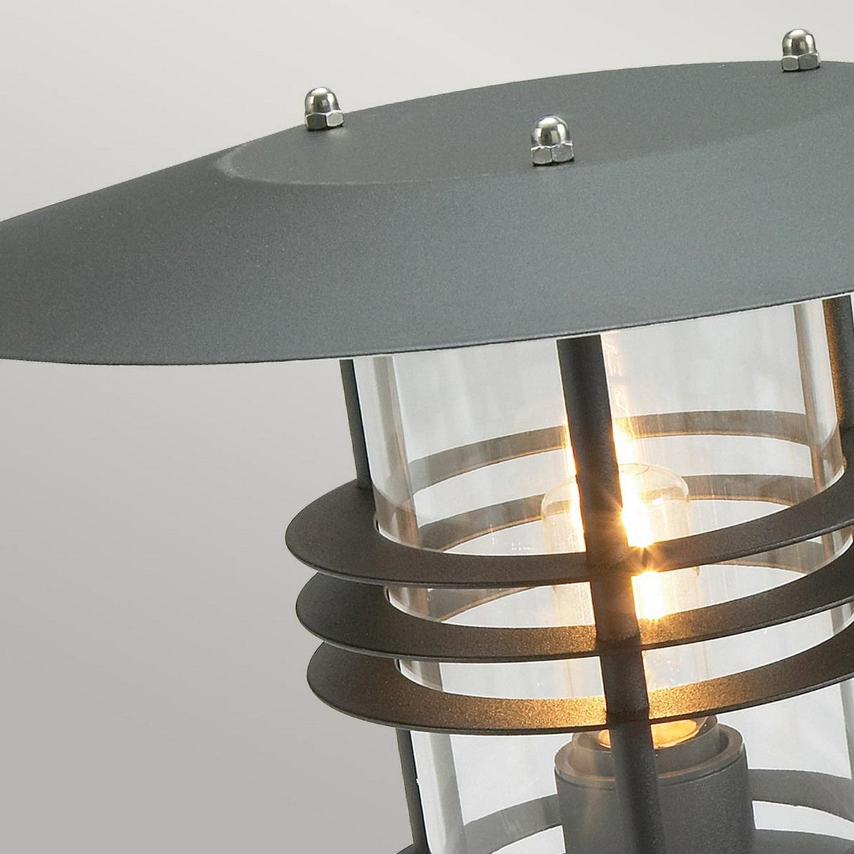A close-up of the Stockholm 1 Light Grande Pedestal in a Scandinavian design showcases its modern style with a cylindrical glass enclosure and concentric metal rings finished in sleek black. The lantern is illuminated, casting a warm glow against a light beige background.