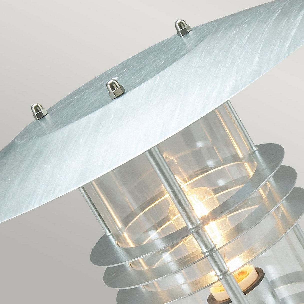 Close-up of the Stockholm 1 Light Grande Pedestal - Silver, a modern outdoor lamp featuring a cylindrical glass casing and multiple metal rings. Crafted from galvanized steel, it emits a warm glow while showcasing its metallic, angled shade secured with small bolts on top. This design embodies Scandinavian elegance.